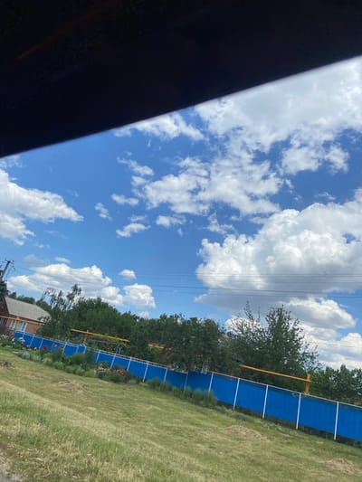 This is a landscape image taken outdoors, with the camera oriented vertically. The sky dominates the upper half of the frame, featuring a clear blue expanse punctuated by fluffy white cumulus clouds. Power lines traverse the sky diagonally from the upper left to the lower right.
The lower half of the image depicts a grassy area with a prominent blue fence running parallel to the right edge. Behind the fence, a line of trees forms a dense green border. A small section of a brick building with a light-colored roof is visible in the lower right corner, near the fence. A yellow structure, possibly part of a fence or pole, is also visible behind the blue fence. The ground in front of the fence is covered in grass, some of which appears dry or sparse. Flowers are visible in a garden bed along the fence line.
The lighting suggests it is daytime, likely a sunny day given the bright sky and well-defined clouds. The scene appears to be a rural or suburban setting, possibly a backyard or park area, in Tagarog, Russia. There is no discernible activity or event taking place, and no visible text. The image evokes a sense of calm and openness due to the expansive sky and greenery.