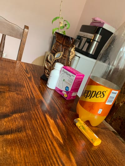 The image depicts a close-up, angled view of a wooden table. On the table, a nearly empty plastic bottle of Schweppes orange soda is prominent. To the left of the bottle, there is a pink and white box labeled "PreMama duo" with some text in Bulgarian, and a white bottle with a white cap. A yellow lighter with "FOOTBALL" printed on it lies in the foreground. In the background, a portion of a wooden chair and a decorative wooden vase with some plant sprigs are visible. A coffee maker and stacked boxes are also partially seen in the background. The lighting suggests an indoor setting, possibly a home. There are no people or explicit actions captured in the image. The location of Krushev Dol, Bulgaria is not visually represented in the image.
