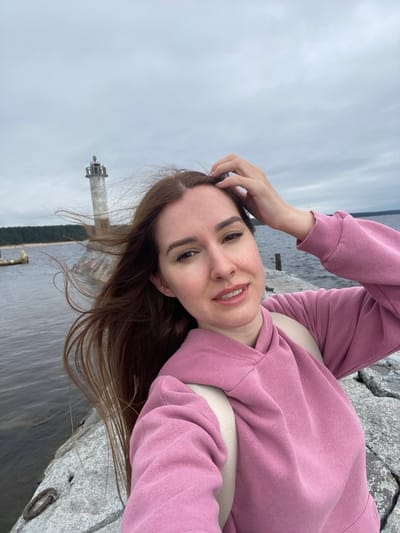 A young woman with long, brown hair, wearing a pink hooded sweatshirt and a backpack, is captured in a selfie. She is standing on a rocky pier next to a body of water. In the background, a lighthouse stands on the opposite shore, with a forest line behind it. The sky is overcast. The location is identified as Моторное, Russia.