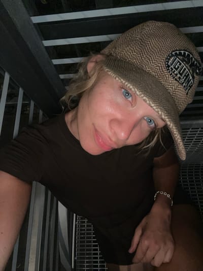 In Ohlstadt, Germany, a young person with blonde hair and striking blue eyes is captured in a close-up, high-angle selfie. They are wearing a brown t-shirt and a patterned brown cap with a dark patch featuring the word "IGUANA" in white lettering. The individual is looking towards the camera with a slight smile, their skin illuminated by ambient light. A bracelet adorns their left wrist. The background suggests an outdoor setting with industrial elements, possibly stairs or a metal structure with horizontal and vertical bars, indicative of a public space or possibly an outdoor staircase in a wooded area given the glimpse of foliage behind the structure. The lighting is dim, contributing to a somewhat moody atmosphere.