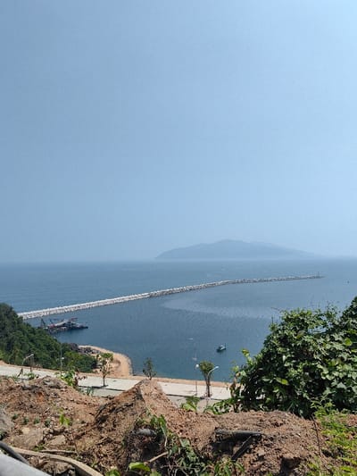 This image presents a high-angle panoramic view of a coastal landscape in Hải Vân Ward, Vietnam, under a clear, light blue sky. The upper third of the frame consists of a pristine, bright blue sky. The middle section is dominated by an expansive, calm dark blue sea.
In the immediate foreground, a steep, reddish-brown dirt embankment with sparse green vegetation and exposed soil occupies the bottom portion, indicating recent land disturbance. Large, dense green foliage is visible on the right edge of this foreground. Below the embankment, a paved, dark grey asphalt road curves along the coastline, featuring multiple white streetlights and small, recently planted trees with light-colored trunks. A narrow strip of light-colored sand or gravel beach separates the road from the sea.
To the left, a verdant, tree-covered hillside slopes down to the water's edge. Extending into the sea from this area is a prominent, long, L-shaped breakwater constructed from numerous light-colored rock or concrete segments. This structure curves gently to the right as it stretches into the bay, protecting an inner harbor area. Moored near the base of the hillside, adjacent to the breakwater, are several dark-colored industrial vessels, possibly barges or dredges. Further out in the bay, towards the center-right, a single small, light-colored boat is visible on the water.
In the distant background, a large, hazy, dark green mountainous landmass or island appears on the horizon, stretching across the right half of the image. There are no discernible people or explicit actions depicted within the scene.