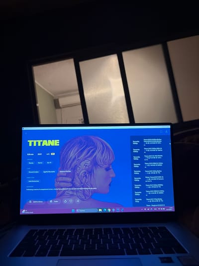 The image displays an open laptop computer in a dimly lit interior setting, identified as being in the city of Karadjovo, Bulgaria. The laptop screen is the primary light source and exhibits a movie information page for "TITANE." The page shows the movie title in yellow, with details including a runtime of "108 min," year "2021," a 6.5 IMDB rating, and genres listed as "Drama," "Horror," and "Sci-Fi." Cast members Vincent Lindon, Agathe Rousselle, and Garance Marillier are listed, along with director Julia Ducournau. A partial summary reads, "Following a series of unexplained crimes, a father is reunited with the son who has been missing for ten years."
The dominant visual on the screen is a stylized profile illustration of a person with short, light-colored hair, large hoop earrings, and intricate, possibly tattooed or augmented, designs around the ear and neck area, set against a vibrant blue background. To the right of this information, a vertical list of downloadable versions of "Titane" is displayed, primarily from "Torrrentio" in 1080p resolution, detailing various file sizes. The Windows taskbar is visible at the bottom of the screen, featuring multiple application icons, a search bar, and the system tray showing the current time as 23:42 and the date as 15.3.2023 (March 15, 2023), with the language set to English.
Above the laptop, a multi-pane window is visible. Through the leftmost pane of this window, part of an adjacent interior room is reflected or visible, revealing light-colored walls, a white ceiling, and a white, horizontally mounted air conditioning unit high on the wall. The overall environment suggests a nighttime setting where the laptop screen provides significant illumination. No individuals are visible in the scene.