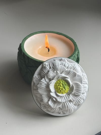 This media file shows a lit candle with a decorative lid. The candle is in a green, textured container that appears to be made of a material resembling leaves. The candle wax is off-white, and a flame is visible, casting a warm glow. The lid is round and white, with a raised, intricate design of a flower with multiple petals and a textured, green center. The lid is partially covering the candle. The background is a plain, light-colored surface, possibly a table or countertop. The lighting suggests it is daytime, and there are no obvious clues about the weather or a specific outdoor location. The overall mood is calm and tranquil. There is no visible text in the image.