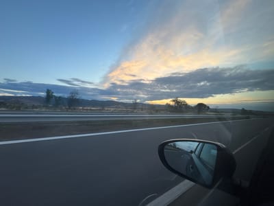 The image depicts a view from inside a moving vehicle, looking out the passenger side window and into the side mirror. The scene is set on a highway with a clear asphalt road and a white line marking the lane. In the distance, rolling hills and sparse vegetation are visible under a twilight sky. The sky is a gradient of blues, with scattered clouds illuminated by the setting sun, casting warm hues of orange and yellow. The side mirror reflects the road ahead, showing the car's perspective and a portion of the sky. The overall impression is one of travel and a serene natural landscape at dusk. The image does not contain any explicit information to pinpoint the location within the city of Kalugerovo, Bulgaria, but it captures a typical highway scene that could be found in many rural or semi-rural areas.