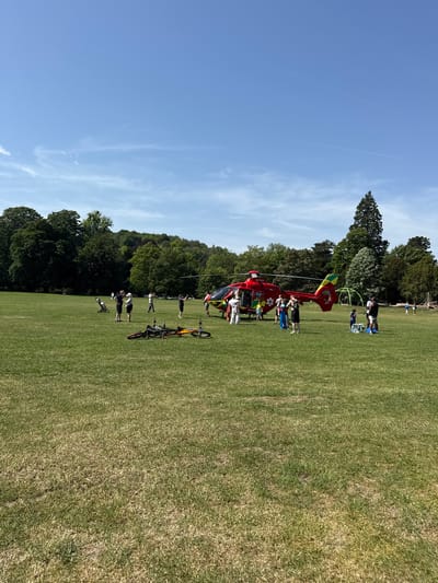 A red and yellow helicopter with the marking "G-TILY" is grounded on a grassy field in High Wycombe, United Kingdom. Multiple individuals are standing around and near the helicopter. In the foreground, there are two bicycles laying on the grass. The background consists of green trees and a blue sky with wispy clouds. Additional individuals can be seen at the far end of the field.
