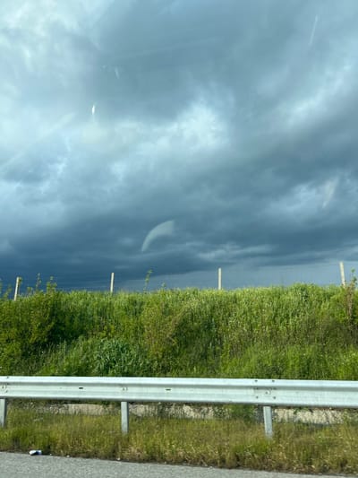 The image shows a view from inside a vehicle, likely on a highway or road. On the right side of the frame, a metal guardrail runs along the road. Beyond the guardrail, there is a grassy verge with dense green vegetation. In the background, there are utility poles and possible a fence. The sky above is overcast with dark, stormy-looking clouds. The reflections in the windshield obstruct the view and limit detailed assessment of the environment. The exact location in the city of Градина, Bulgaria, cannot be determined from the provided image.