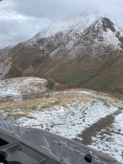 The image depicts a snow-dusted mountainous landscape in Juta, Georgia, viewed from inside a vehicle, likely an off-road utility vehicle, with its dashboard and windshield partially visible in the lower frame. The sky is overcast and uniformly grey. In the midground, rolling hills are partially covered with patches of white snow and dry, brown-yellow grass. A narrow dirt road or path winds through this terrain. A small, white, single-story building with a dark roof is situated on a lower slope on the left, near a visible fence line. To the right of this building, a dark brown horse grazes in the snow-dusted grass. Further into the midground, a solitary deciduous tree with green foliage stands on a small rise, with green support poles nearby. Distant mountains rise steeply, their upper slopes covered with fresh snow, while lower sections reveal brown, sparse vegetation. Another dirt path is visible at the base of the distant mountains. No people are visible. The scene suggests a cold, early winter or late autumn environment.