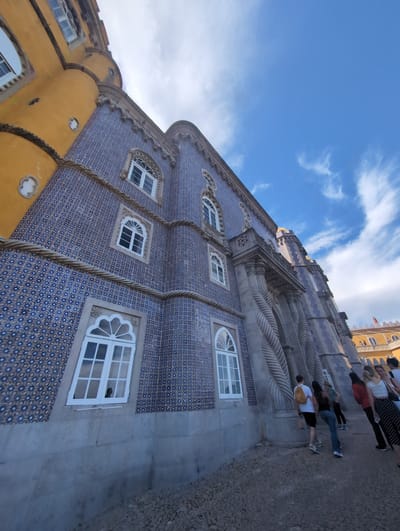 Captured from a low-angle perspective, the image features a section of the Pena Palace located in Sintra, Portugal. The architectural facade includes blue tilework with rows of arched windows with white trim. A yellow, rounded corner section of the palace is also visible. In the background, the light blue sky is dotted with white clouds. A group of tourists can be seen walking in front of the palace. The image is rotated counter-clockwise.