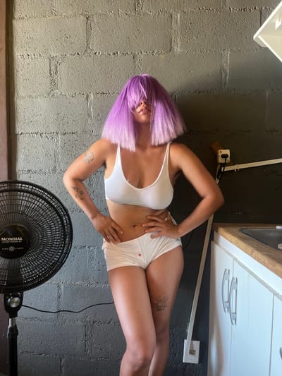 An adult individual stands in an indoor setting within Arraial Do Cabo, Brazil. The individual wears a short, shaggy, bright purple wig that obscures the upper face, revealing only the lower lip. They are clad in a white sports bra and white boy-short style underwear, which has a small red mark or design on the front left. Visible tattoos include an faded design on the left bicep, two small linear tattoos on the left forearm, and a stylized sun or burst tattoo on the right thigh. The individual stands with hands on hips, striking a pose. To the left, a black "MONDIAL" brand standing electric fan is positioned. The background features a dark gray concrete block wall. On the right, a kitchen area is visible, comprising a light-colored wooden countertop with a metallic sink, situated above white cabinets equipped with silver handles. A white wall-mounted power outlet with a visible plug is located near the bottom right corner of the frame, against the gray wall, with a white electrical conduit extending horizontally above it.