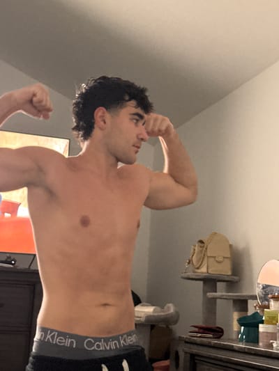 A young, shirtless man with dark, curly hair is flexing his biceps in a dimly lit room. He is wearing Calvin Klein underwear. The background shows a dresser, a television, and a shelf with a purse, a mirror, and various containers. The scene appears to be indoors, likely a bedroom, in Bellmore, United States of America.