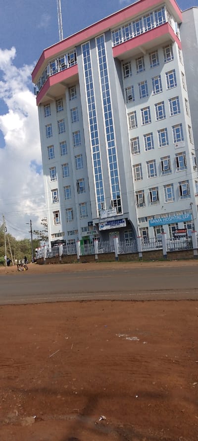 A tall building dominates the frame, featuring a light blue facade with white-framed windows and a pink-accented top floor. Signs at the base of the building identify it as "BAMA HOSPITAL LLP" and "MEDZORE HEALTHCARE." A street runs in front of the building, dividing it from a dirt patch foreground. A few people can be seen on the street, and power lines stretch across the sky in the background. Clouds are visible in the blue sky.