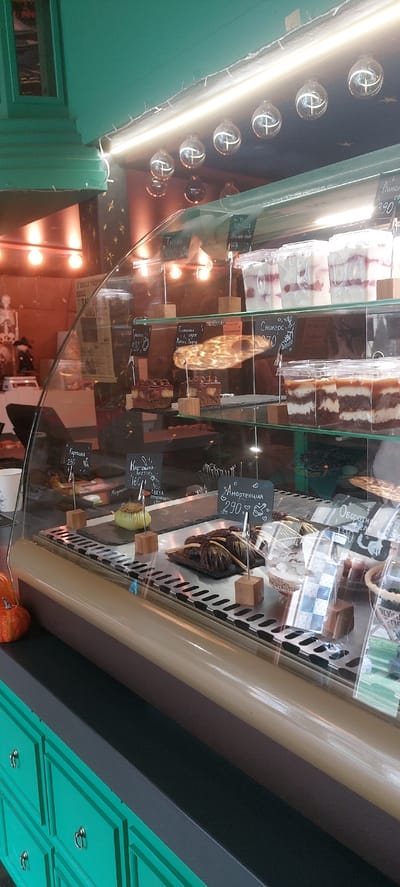 This image depicts a refrigerated display case filled with various desserts, likely in a cafe or bakery. The case is illuminated from above by string lights and several round, decorative light bulbs. Inside the case, desserts are arranged on glass shelves. Some items appear to be individual pastries, while others are in clear plastic containers. Small chalkboards with handwritten labels and prices are placed in front of many of the items, indicating a retail or service environment.
On the top shelf, several transparent containers hold layered desserts, possibly parfaits or trifles, with visible fruit and cream. Below this, a dark brown cake with chocolate toppings is displayed on a lower shelf, alongside other similar pastries. Further down, a unique green dessert shaped like a pear is presented, with a small chalkboard labeling it "Картошка-пюре" (mashed potatoes) and "160". Next to it, there's a tray of dark, cone-shaped chocolates that resemble brooms, labeled "Амортенация 290". Another section of the case shows layered chocolate and cream desserts in clear containers. On the far right, a partially visible dessert is in a patterned container.
In the background, behind the display case, there are glimpses of a wall decorated with what appear to be star shapes and some posters. A miniature skeleton figure and a witch figurine are also visible, suggesting a possible theme or seasonal decoration. The exterior of the display case is encased in glass, reflecting some of the interior items and the lighting. The base of the display unit has green drawers with ornate handles.
The specific location of this scene within the city of Berdsk municipality, Russia, cannot be determined from the image alone. The presence of Cyrillic script on the price tags strongly suggests a Russian-speaking region.