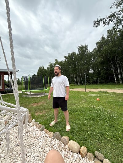 A man with a beard, wearing a white t-shirt and black shorts, stands in a grassy yard under a cloudy sky. He is looking up and appears to be shouting. In the background, there is a trampoline, a volleyball net, and a line of trees. To the left of the frame, a white swing is partially visible. The location is identified as Ryzhkovo, Russia.