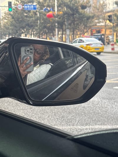 The image is taken from inside a vehicle, focusing on its passenger-side exterior mirror and the street scene beyond. Reflected in the mirror is an individual, presenting with dark skin, wearing glasses, and having light-colored, curly hair. This person is holding a grey smartphone (identified by its multi-lens camera array and a circular magnetic accessory/ring holder) in their right hand, pointed towards themselves, indicative of taking a selfie. They are wearing a white quilted jacket or vest over a dark garment. The vehicle's interior visible in the reflection includes a white quilted door panel.

Outside the mirror, an urban street environment is visible under daylight conditions. A green traffic light is illuminated in the upper left quadrant. A blue road sign displaying Chinese characters, including "黄山路" (Huangshan Road), confirms the location as Huanggu District, Shenyang, China. The street is lined with trees, currently appearing bare-branched. A red, spherical object, possibly a decorative lantern, is suspended overhead from a pole. A two-tone white and yellow sedan, equipped with a roof-mounted light bar, consistent with a taxi or an official vehicle, is observed in the middle distance, traveling away from the viewer. Red and white striped traffic bollards mark the roadside on the right. The road surface consists of grey asphalt.