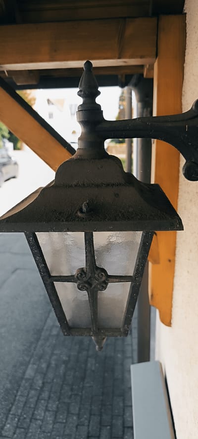 An exterior shot, captured in Schwaikheim, Germany, focuses on a black, ornate, old-fashioned lantern attached to the side of a building. The lantern features a dark metal frame with clear, textured glass panels. It is suspended from a decorative metal bracket that is bolted into a wooden beam and the exterior wall. The building has a light-colored, textured facade. To the left of the lantern, a diagonal wooden beam, stained a warm, light brown, is visible. In the background, a paved area, likely a street or walkway, with a grey, brick-like pattern is out of focus. A blurred car and some foliage are also discernible in the distance. The overall impression is one of a vintage exterior fixture in a residential or commercial setting.