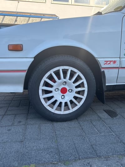A close-up, side-angle shot depicts the front tire and fender of a white car, parked on a gray brick surface. The tire features a white, multi-spoked rim with a red center cap. The tire is marked with "185/55R14 80H" and "MADE IN CHINA,". "スズキ" (Suzuki) is printed in red on the fender, along with a red stripe. An orange side marker light is visible below the fender. The background suggests an urban environment. The car and the brick pavement are located in Fulda, Germany.