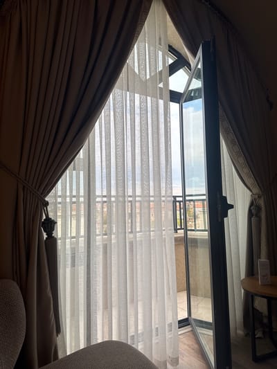 The image shows an interior view looking out through an open window, likely in Ürgüp, Turkey, although specific landmarks are not visible. The window frame is dark grey or black. Sheer white blinds or shades partially obscure the view. Thick brown drapes are drawn to the sides of the window, tied back with a decorative tassel. A small section of what appears to be a chair is visible in the lower right of the frame, covered in a light grey fabric. A small table with a wooden top and metal frame can be seen in the upper right. The view through the window is of a building and sky.