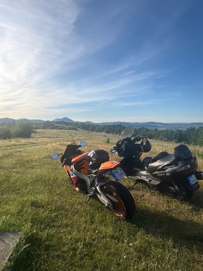 Two motorcycles are parked on a grassy hill overlooking a valley. The motorcycle in the foreground is an orange and white sportbike, and the motorcycle behind it is a dark-colored scooter. Both motorcycles have license plates visible. The background features rolling hills and mountains under a blue sky with wispy clouds. The scene appears to be in a rural area.
