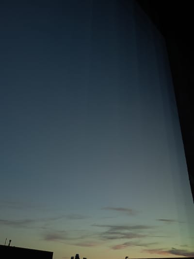 The image depicts a twilight sky over Peetri alevik, Estonia. The sky transitions from a deep navy blue at the top to lighter shades of blue and hints of pink and yellow near the horizon, suggesting the sun has recently set. Wispy clouds are visible in the lower portion of the sky. Silhouetted against the horizon are the tops of buildings, including what appears to be a chimney or vent pipe on a darker structure in the lower left corner. There are no people or active events visible in the scene. The overall impression is one of a quiet, evening cityscape.