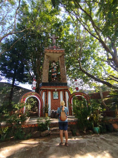 A woman stands before a religious shrine in An Phu Ward, Vietnam. The shrine is decorated with arches, red and white stripes, and a statue of a woman and child. The shrine is partially shaded by trees. The woman is wearing a light blue shirt, blue shorts, sandals, a hat, and a backpack. She is facing the shrine. The ground is dirt. There are plants and trees surrounding the shrine.