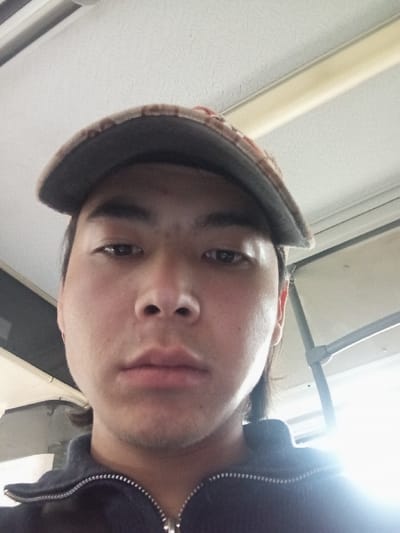 A close-up selfie captures a male individual with an East Asian or Central Asian phenotype, looking directly at the camera with a neutral expression. He wears a camouflage-patterned baseball cap, positioned high on his forehead, and a dark, possibly navy or black, zip-up jacket with a silver zipper. Dark hair is visible extending from under the cap. The background features a light-colored, textured ceiling, consistent with an indoor environment or vehicle interior. A linear fluorescent light fixture runs along the ceiling. A structural bar with a gray joint is present in the upper right. A strong light source on the right side creates significant glare. The image provides no discernible visual cues to confirm a specific location within Zangiata, Uzbekistan.
