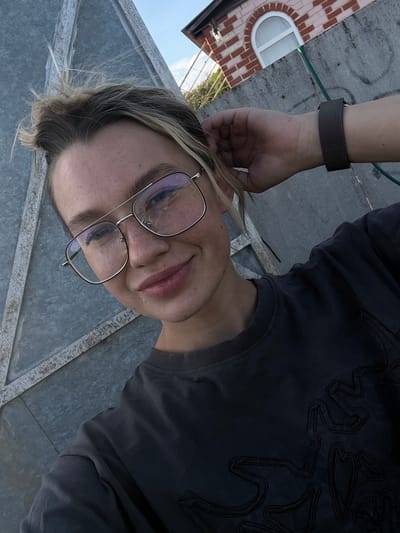 A young person with blonde hair and glasses is looking directly into the camera. The person is wearing a dark grey shirt with a decorative pattern. A metal watchband is visible on the person's wrist. In the background, there is a brick building with a curved window, suggesting an urban setting. The setting is possibly within the city of Слобода, Russia, based on the location information.
