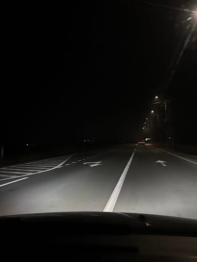 The image depicts a nighttime view from the interior of a vehicle, looking forward onto a two-lane asphalt road in Vama Veche, Romania. The lower portion of the image shows the dashboard or hood of the originating vehicle, illuminated by its own headlights, which cast light directly onto the road surface.
The road features clear white markings: a solid center line separating traffic directions, a straight-ahead arrow in the right lane, and a left-turn or straight-ahead arrow with a diagonal striped area in the left lane, indicating a potential merge or turning bay. Along the right side of the road, a series of evenly spaced streetlights mounted on wooden utility poles illuminate the immediate vicinity, casting distinct pools of light.
In the distance, a single vehicle is observed traveling in the same direction on the right lane, identifiable by its illuminated red taillights. The environment beyond the illuminated road and immediate streetlight range is enveloped in deep darkness, with no discernible structures or clear natural features visible. No individuals are present in the frame.