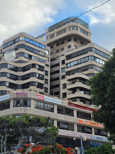 The image shows a modern, multi-story building with a distinctive wavy facade in Santa Cruz de Tenerife, Spain. The building has many windows and is adorned with several signs, including "Santander," "ENIC ESCUELA DE NEGOCIOS," "GRUPO CAMPER," and "RACE." Trees and vibrant red flowers are visible in the foreground, suggesting a pleasant outdoor setting. The sky is partly cloudy, indicating daytime. The orientation of the image is portrait, and the building is viewed from a low angle looking upwards. The overall impression is of an urban environment with commercial and educational establishments.