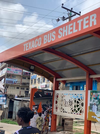 The image shows a bus shelter in Ondo, Nigeria. The shelter has an orange roof with the words "TEXACO BUS SHELTER" written in white letters. The shelter is supported by red metal beams. Underneath the shelter, there is a metal gate with white cut-out designs, and two posters. The posters have various advertisements in a local language, with the words "ONDO IS LUCKY" and "ODA TI WA 2024" visible. Behind the shelter, there are residential buildings with visible signs, and a blue building to the right. A young woman wearing a purple and white shirt is standing to the left of the shelter. A boy in a black shirt is standing near her. The environment is typical of a developing city. The sky is partly cloudy. There are power lines with numerous electrical poles. The image was taken on a sunny day.