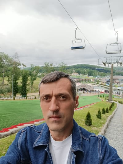 A selfie-style portrait captures a middle-aged male with short, dark hair exhibiting visible grey at the temples, looking directly at the camera with a neutral expression. He is wearing a blue denim jacket over a white crew-neck t-shirt. The background depicts an outdoor recreational area in Vynnyky, Ukraine, under an overcast sky. To the man's left, a wide expanse of bright green artificial turf, marked with red and white borders, extends, resembling a dry ski slope or summer tubing track. Beyond this, a treeline and a light pole are visible. To the right, a stone pathway lined with small, conical evergreen shrubs ascends. Overhead and to the right, multiple empty ski lift chairs are suspended from cables, supported by a prominent metal ski lift tower. In the mid-distance, a complex of buildings, some featuring thatched roofs, is visible adjacent to a body of water. Distant rolling, forested hills complete the horizon.