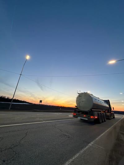 The image depicts a tanker truck driving on a paved road at dusk. The road appears to be a highway near Kuregovo, Russia, given the landscape and street lighting. A dark wooded area is visible alongside the road where the sun sets. The sky transitions from dark blue to orange near the horizon. Street lights illuminate the road, and the tanker truck's tail lights are visible. No other vehicles or people are evident in the frame.