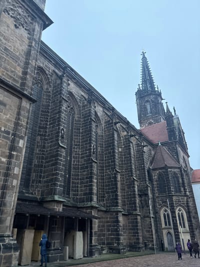 This image features a majestic, historic church or cathedral, viewed from a low angle looking upwards, likely depicting Meissen Cathedral in Germany. The primary subject is the colossal stone edifice, constructed from dark, weathered blocks, showcasing intricate Gothic architecture.
The main body of the church is characterized by robust buttresses and ornate window frames with delicate tracery patterns. Its side walls rise steeply, punctuated by rhythmic architectural elements. A reddish-brown roof section is visible where the main building transitions to a tower.
Dominating the background against a light, overcast sky is a very tall, slender spire. This spire is richly detailed with multiple tiers of pointed pinnacles and complex ornamentation, culminating in a cross at its apex. Along the roofline of the church's main structure, several dark, stylized figures, possibly gargoyles or decorative elements, add to its Gothic character.
The sky is a uniform, pale grey-blue, indicating an overcast or cloudy daytime setting without direct sunlight. In the lower right corner, several small figures are visible. One person, wearing a blue jacket, stands near large, light-colored stone blocks at the base of the church. Other figures are further in the background on what appears to be a paved area. Their attire suggests cool weather. No discernible text is visible within the image.