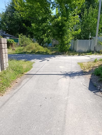 A paved road in Lenghir, Kazakhstan branches into two paths. To the left, a brick pillar stands by the roadside, with green grass and vegetation growing alongside. Beyond that, a corrugated metal fence encloses an area with trees and a building with a dark roof. To the right, the road continues past sparse vegetation and rocks, leading towards a similar fence and a utility pole. The area is bathed in bright sunlight, casting shadows from the trees onto the road.