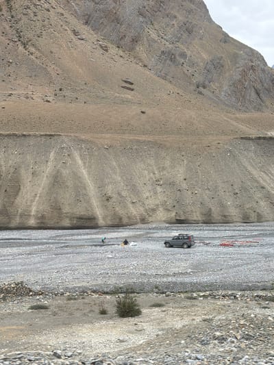 In Zangla, India, a grey SUV is parked in a rocky riverbed. Two individuals, one wearing a turquoise shirt and the other a black shirt, are in the distance, appearing to work near the river. The riverbed is covered in grey pebbles and stones. Behind the riverbed, a steep, barren hillside rises, characterized by layers of rock and soil. The sky is overcast.