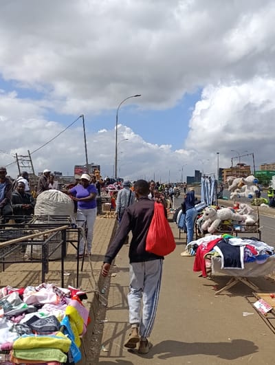 The image captures a bustling open-air market scene, likely situated along a roadside or bridge in Kiuu ward, Kenya. The setting is vibrant with activity under a partly cloudy sky, showing patches of blue.
In the foreground, a man wearing a dark long-sleeved top and light grey sweatpants with blue stripes walks away from the camera, carrying a bright red bag over his left shoulder. His shadow is cast on the ground beside him. To his left, colorful piles of clothing and textiles are displayed directly on the ground next to a raised curb, indicating items for sale. Behind these, several empty metal market stalls or frames are visible.
Further into the scene, a woman in a purple top and light pants stands near a large, bulging white sack, potentially containing more goods. On the right side of the frame, another makeshift stall displays various clothing items on a table, alongside several large stuffed animals, including white bears and a yellow lion. Numerous other people are scattered throughout the background, appearing as vendors, shoppers, or pedestrians moving through the market.
The scene depicts a lively marketplace where various goods, primarily clothing and toys, are openly displayed. It is daytime, evidenced by the bright sunlight and visible shadows. The environment includes streetlights lining the road, and an elevated structure or bridge spans across the background. Power lines and a rustic-looking utility pole with a ladder-like attachment are visible on the left side of the upper frame. A distant billboard in the mid-ground shows partially legible text that appears to read "Opti-Rana". The overall atmosphere is active and commercial, characteristic of a busy local market.