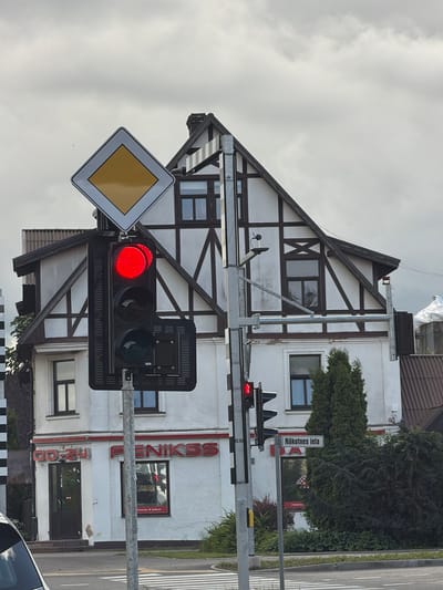 The image shows a street view in the city of Ķekava, Latvia. In the foreground, a traffic light shows a red signal for vehicles and a red pedestrian signal. A diamond-shaped sign with a yellow center and white border, indicating a main road, is mounted above the traffic light. To the right of the traffic light, a street sign reads "Nākotnes iela". In the background, a building with a distinct half-timbered facade and dark trim is visible. The building has multiple windows and a sloped roof. Below the half-timbered section, the building is painted white and features store signage, including "00-24" and "FENIKSS". Several trees and bushes are present in the scene. The sky is overcast.