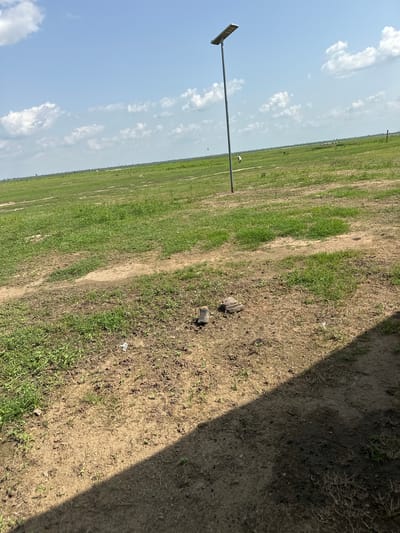 A wide shot captures a vast, green, grassy plain under a bright, cloudy blue sky. A tall, slender pole with a solar panel is prominently positioned on the left side of the frame, with its shadow stretching across the foreground. The ground in the foreground is a mix of dry, sandy soil and patches of short green grass. In the middle ground, two small, brown objects that appear to be pieces of wood or rocks are visible. The horizon is flat, with more green fields stretching out, dotted with a few distant figures that could be animals or small structures. The overall scene depicts an open, rural landscape in Lamurde, Nigeria.