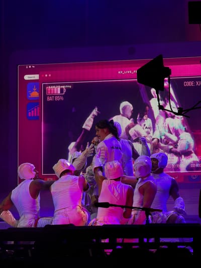 The image captures a live performance, likely a concert or stage show, bathed in purple and pink stage lighting. In the foreground, several performers are dressed in matching white outfits, including white sleeveless tops, white pants, and white helmets or head coverings that give them a futuristic or stylized appearance. One performer in the center, a woman wearing a white jacket with the word "HUMAN" visible on it, is singing into a microphone. She has dark hair pulled back and is looking towards the left of the frame. Other performers are arranged around her, some facing her, others with their backs to the camera.
In the background, a large screen displays graphics that resemble a digital interface. On the screen, there's a section showing "2,500,213 watching" next to a battery icon indicating "BAT 85%". Other text elements like "KP_LIVE_FE" and "CODE: XJ-" are also visible. Further down the screen, text reads "WE A LABEL" and "REC 00:15:32 ISO 800". The overall atmosphere is energetic and theatrical, with a clear focus on the performers and the visual presentation. The location is not specified in the image itself.