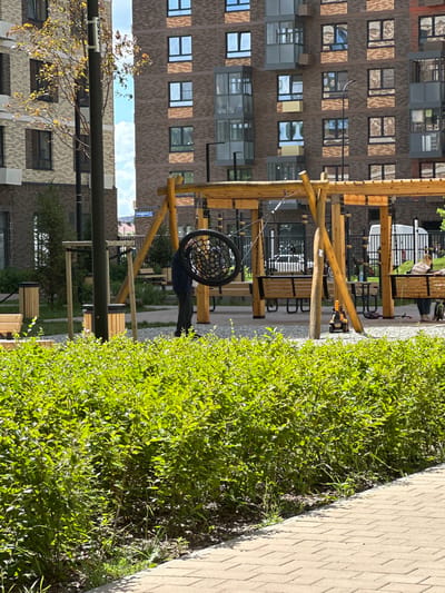In Znamya Oktyabrya, Russia, a contemporary residential courtyard is captured on a sunny day. The scene features a multi-story brick building with numerous windows and balconies, indicating modern architecture. In the foreground, lush green bushes partially obscure a paved walkway and a gravel area. A wooden playground structure with a tire swing is central, with a person dressed in dark clothing adjusting the swing. Several wooden benches are positioned beneath the structure. A small yellow toy excavator and other children's toys are visible on the ground. A dark vertical pole, possibly a lamppost or utility pole, stands to the left of the playground. The sky is a clear blue with some clouds.