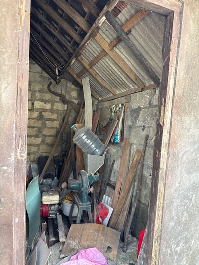 The image depicts a cluttered storage area viewed from above, likely within a building in Pejukutan, Indonesia. The space is filled with construction debris, including wooden planks, corrugated metal roofing, concrete blocks, and other unidentified materials. A water jug is visible, along with a small engine and various containers. The environment appears to be poorly maintained and possibly abandoned. No people are visible in the image.