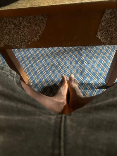 The image presents a close-up, downward-angled view, primarily focusing on a pair of bare human feet resting on a patterned textile surface. The feet appear to belong to an individual with a medium to dark skin tone. One foot, positioned towards the front-right of the frame, is largely visible from the heel to the toes, with the arch and top of the foot prominently displayed. The other foot is partially obscured, positioned slightly behind and above the first, with its toes and a portion of the top visible, seemingly resting gently on or very close to the other foot.

The surface beneath the feet is a rug or carpet featuring a repetitive geometric pattern. This pattern consists of small, alternating squares or diamonds in shades of blue and a light brown or beige, creating a tessellated, houndstooth-like design across the floor.

To the left side of the image, a vertical wooden structure is visible, likely a leg or frame of a piece of furniture such as a chair or table. This wood has a warm, reddish-brown tone. Attached to its inner surface is a lighter, granular material, speckled grey and white, reminiscent of granite or a coarse laminate. This material shows signs of wear, with several sections appearing chipped, peeling, or unevenly applied, revealing glimpses of the underlying wood.

On the far right of the frame, a dark, textured fabric element occupies a significant portion of the foreground. This fabric, likely part of clothing such as trousers (possibly denim due to its texture and visible seams), partially obstructs the view of the feet and the patterned floor, creating a soft, out-of-focus blur.

The lighting in the scene is soft and diffused, casting gentle shadows that define the contours of the feet and the various textures present. The overall perspective suggests an intimate, casual shot, possibly taken by someone looking down at their own feet while seated or resting.