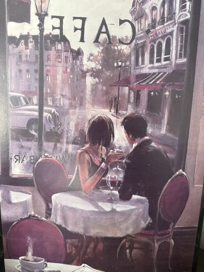 This is a painting of a romantic scene in a European-style cafe, possibly in Paris, France, or a similar city with European architecture. The cafe appears to be situated on a street with buildings featuring balconies and awnings. Through the window, a classic car is visible. Inside the cafe, a couple is seated at a round table covered with a white tablecloth. They are enjoying drinks, with wine glasses on the table. The woman is wearing a sleeveless dress and jewelry, while the man is in a suit jacket. The cafe interior is dimly lit, with ornate chairs and a lamp post visible outside. The overall atmosphere suggests a sophisticated and intimate setting.