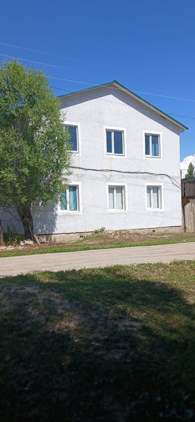 A two-story white stucco house with a green roof is situated in a rural area. The house has six windows on its front facade, three on the upper level and three on the lower level. A large, leafy green tree stands to the left of the house, casting a shadow on the building. A dirt road runs in front of the house, bordered by patchy grass and dirt. In the background, a wooden fence and some distant trees are visible under a clear blue sky. The image was taken in Mihaylovka, Kyrgyzstan.