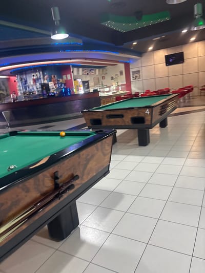 A sideways, slightly high-angle indoor shot shows two pool tables, one with cue sticks, in what appears to be an entertainment establishment, possibly a bar or club, in Gioia Tauro, Italy.
The pool tables are positioned lengthwise from bottom-right to top-left. The lower table has a green felt surface, one visible yellow ball, and two cue sticks resting on its edge. The upper table is marked with the number "4". Both tables are dark brown with black trim.
The floor is covered in white square tiles with light gray grout. Behind the tables, there's a bar area or seating area with red chairs. Further in the background, there's a brightly lit store or bar with various products and displays.
On the left, there is blue lighting. A TV screen is visible on the wall. The overall lighting is bright and artificial. The environment appears to be indoors.