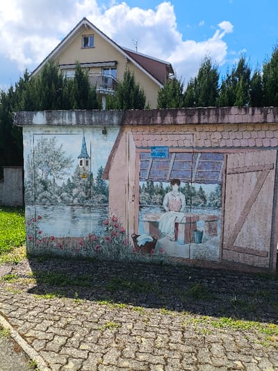 A rectangular, low-rise concrete structure, located in Oberschaeffolsheim, France, displays a multi-panel mural under a clear blue sky with scattered white clouds. The structure is positioned on a grey paver surface, with sparse grass growing between the pavers, and abuts a grassy area to its left. The mural's left panel features a painted landscape scene depicting a body of water, lush green trees, and a distant church spire; pink flowers are painted along the bottom edge of this panel. A yellow triangular warning label with an exclamation mark is affixed to the painted surface on this section. The right panel of the mural depicts a female figure in an apron over a dress, standing behind a stone wash tub containing white laundry. A dark basket filled with fabric and a blue bucket are painted on the ground beside the wash tub. A blue rectangular sign with white text is painted above the figure. Behind the structure, a dense line of tall, dark green coniferous trees rises, partially obscuring a two-story residential building. This building features a light-colored facade, a reddish-brown pitched roof with an antenna, an attic window, and a second-story balcony with a metal railing. Sunlight illuminates the scene, casting shadows in the foreground.