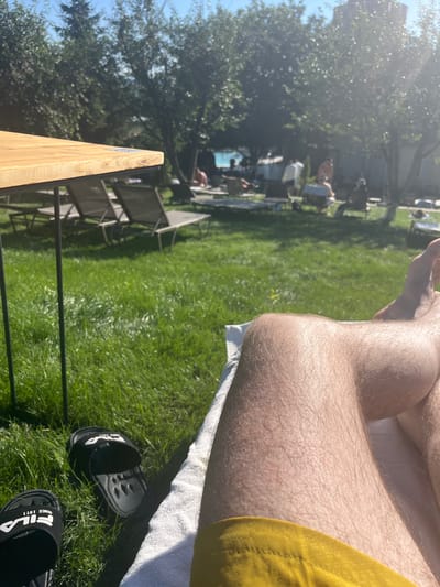The image depicts a sunny outdoor scene with a focus on a person's legs and feet resting on a white towel. In the foreground, a pair of black Fila slides are visible on green grass. The background shows a grassy area with several lounge chairs, some occupied by people who are out of focus. Trees with dense foliage are scattered throughout the area, and the sun is casting dappled light. A wooden table with metal legs is positioned to the left of the frame. The overall impression is one of relaxation in an outdoor recreational space, possibly a resort or park area, given the presence of lounge chairs and people enjoying the sun. The mention of Stăuceni, Moldova, suggests this is a specific location within that city.