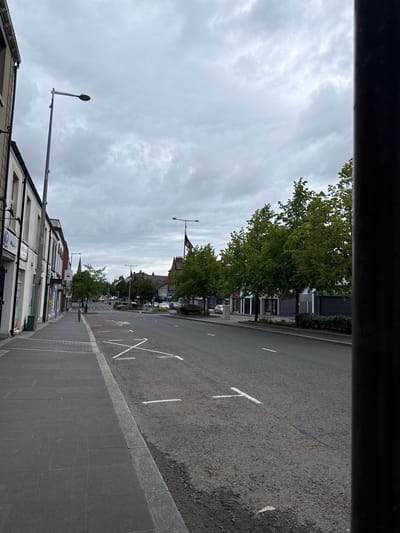 The image shows a street view in Lurgan, United Kingdom. The perspective is from behind a dark, possibly metal, structure at the top of the frame, looking down the street. The street has a gray sidewalk and a paved road with white road markings. Buildings line both sides of the street, varying in height and architecture. Streetlights are visible on the left. Trees line the street on the right. In the distance, there is a building with a steeple and what looks like a flag. The sky is overcast with heavy clouds. There are some vehicles visible in the distance.