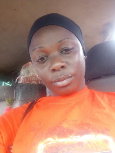 The image presents a close-up portrait of a dark-skinned adult female, captured inside a vehicle in the city of Tataura, Nigeria. She is centrally positioned, facing the camera with a neutral expression. She wears a black head covering and an orange short-sleeved top adorned with a prominent, bright graphic design across the chest, resembling fire or glowing energy. A thin, dark strap from an unseen bag crosses her right shoulder. The background reveals the interior of the vehicle, characterized by a light brown ceiling. To the subject's left, a light-colored sack, partially visible and marked with text, including the word "CEMENT," rests behind her. Below the sack, a dark, textured, possibly mesh-like object is present, with faint green foliage visible behind it. A bright, localized light source or reflection creates a glare in the upper right portion of the background. The scene is static, depicting the individual in a posed stance within the vehicle's confined environment.