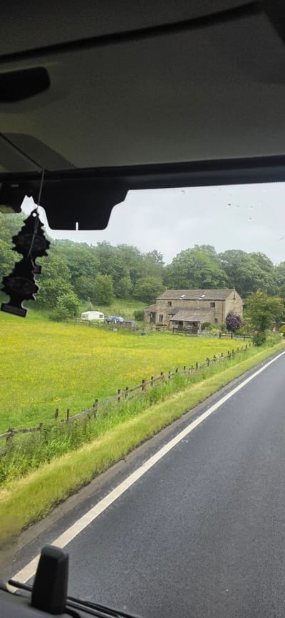 The image depicts a rural scene, likely in or around Barnsley, United Kingdom, as suggested by the context. The viewpoint is from inside a vehicle, looking out through the windshield. In the foreground, a black car air freshener shaped like a pine tree hangs from the rearview mirror. A paved road with a white line runs along the right side of the image, bordered by a grassy verge.

In the mid-ground, a wooden fence runs horizontally across a vibrant green field dotted with yellow wildflowers. Beyond the field, a stone building, possibly a farmhouse or barn conversion, is visible. The building has a tiled roof and several windows. To the left of the building, a white caravan and a dark-colored SUV are parked in the yard. The background consists of a dense line of lush green trees, indicating a wooded area. The sky is overcast, suggesting a cloudy day. No people are visible in the frame.