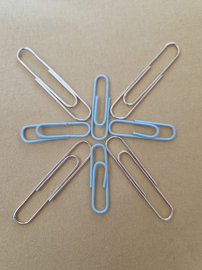 A top-down shot shows a collection of paper clips arranged in a symmetrical pattern on a textured, light brown surface. Five silver-colored paper clips are positioned with their loops facing outwards, forming the points of a star or asterisk. Four of these silver clips are oriented along the four cardinal directions, while one is placed directly between the top and right clips. In the center of the arrangement, four light blue paper clips are clustered. Two blue clips are horizontal, one above the other, and two are vertical, one to the left and one to the right of the horizontal pair. The surface beneath the paper clips appears to be a woven fabric. There is no indication of the location in Fresnes, France, or any people present in the image.